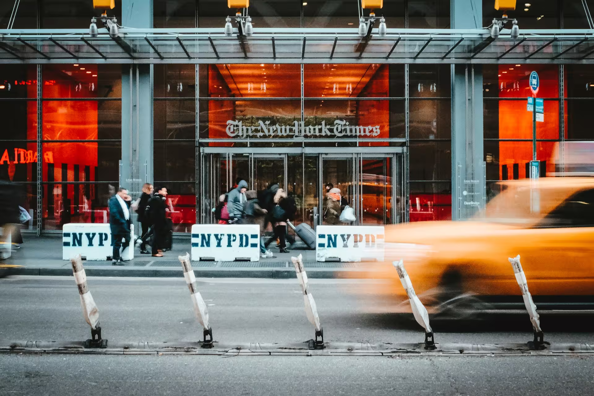 Entrance to The New York Times building, with pedestrians and a passing taxi.